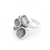 A silver ring featuring four labradorite stones of varying shapes and sizes. The stones include a rectangular, two circular, and an oval-shaped labradorite, all set in a modern, artistic design. The labradorite stones display a subtle play of colours, characteristic of the gemstone.