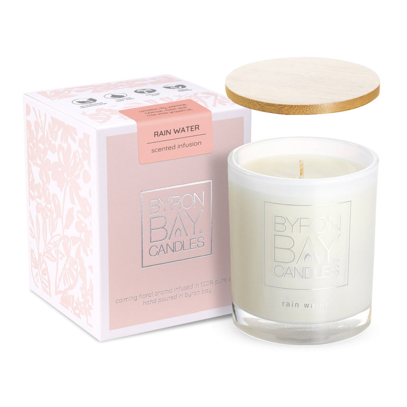 A white candle in a glass container with the label 'BYRON BAY CANDLES' and the scent 'Rain Water' is displayed next to its packaging. The packaging is pink with floral designs and features a label that reads 'RAIN WATER scented infusion.' A wooden lid rests on top of the box. This image shows a beautifully presented Rain Water scented candle from Byron Bay Candles, emphasizing its elegant design and refreshing fragrance.