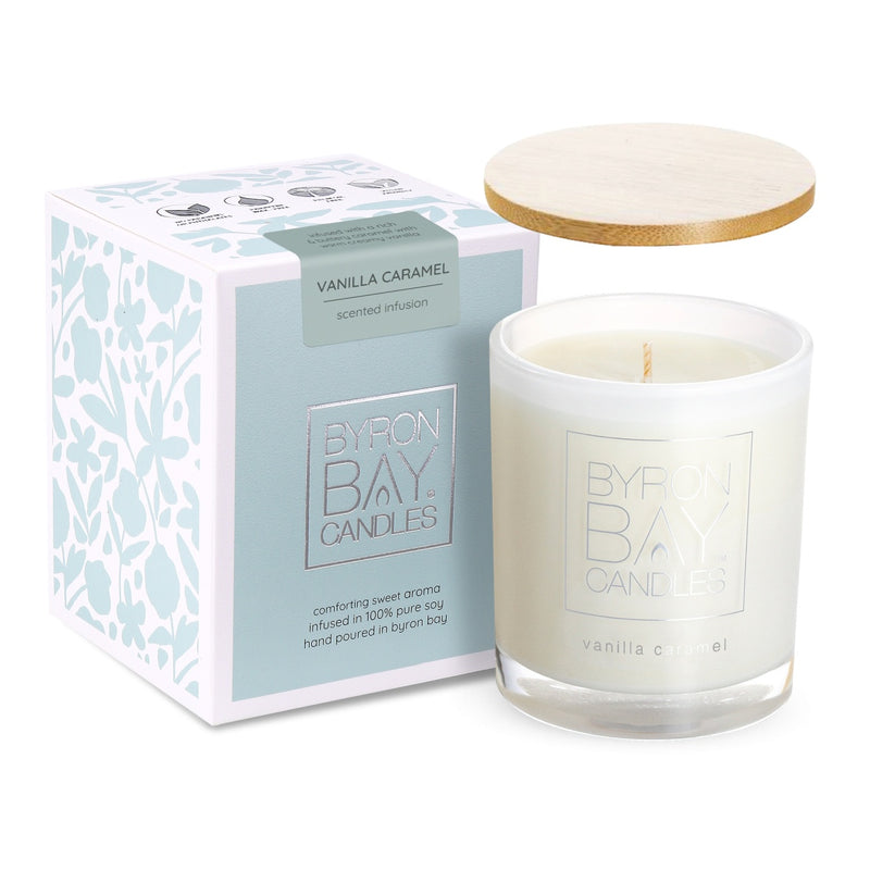 A white candle in a frosted glass container with the label 'BYRON BAY CANDLES' and 'vanilla caramel' is shown next to its packaging. The packaging is a light blue box with white floral patterns and a label that reads 'VANILLA CARAMEL scented infusion.' The box also has text that says 'comforting sweet aroma infused in 100% pure soy hand poured in Byron Bay.' A wooden lid is placed on top of the box.