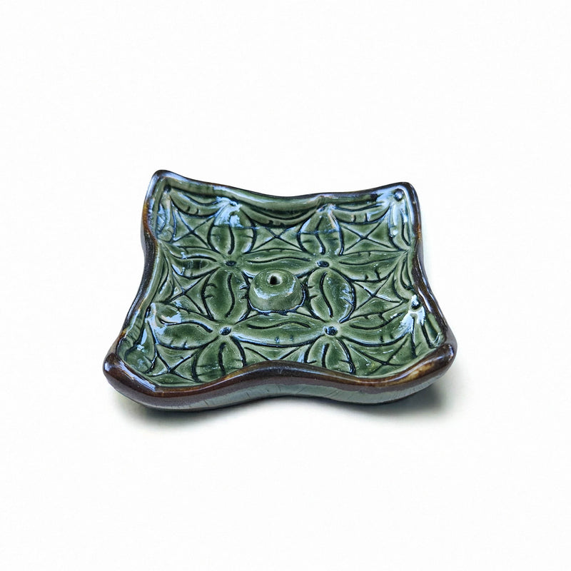 A beautiful green ceramic incense holder with a wavy rectangular design featuring intricate floral patterns. The holder has raised edges with a warm, brownish tint and a small hole in the centre for holding an incense stick. The blend of colours and textures showcases detailed craftsmanship, making this piece both functional and decorative.