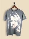 A slate green T-shirt featuring a bold, high-contrast stencil-style portrait of  Kurt Cobain with long hair in an off-white print. 