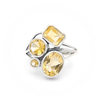 The image shows a silver ring adorned with four citrine gemstones. The gemstones are of different shapes: a rectangular cut, two round cuts, and an oval cut. The arrangement and variety of gemstone shapes make the ring visually interesting and unique.