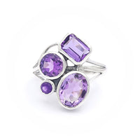 A silver ring featuring four amethyst gemstones of varying shapes and sizes. The gemstones include a rectangular cut, a round cut, an oval cut, and a small circular cut, all set in a modern, artistic design.