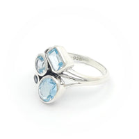 The image shows a silver ring adorned with four blue topaz gemstones. The gemstones are of different shapes: a rectangular cut, two round cuts, and an oval cut. The ring has a modern and artistic design with a split shank band, making it visually interesting and unique.