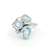 The image shows a silver ring adorned with four blue topaz gemstones. The gemstones are of different shapes: a rectangular cut, two round cuts, and an oval cut. The ring has a modern and artistic design with a split shank band, making it visually interesting and unique.