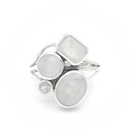 A silver ring featuring multiple moonstones in various shapes and sizes. The design includes a rectangular moonstone, two round moonstones, an oval moonstone, and a small circular moonstone, all set in an elegant silver band. The moonstones have a captivating, milky translucence, adding a touch of ethereal beauty to the ring.