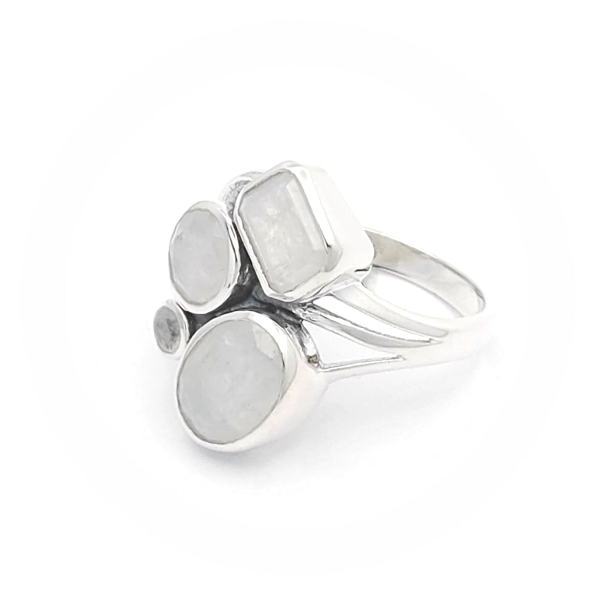 A silver ring featuring multiple moonstones in various shapes and sizes. The design includes a rectangular moonstone, two round moonstones, an oval moonstone, and a small circular moonstone, all set in an elegant silver band. The moonstones have a captivating, milky translucence, adding a touch of ethereal beauty to the ring.