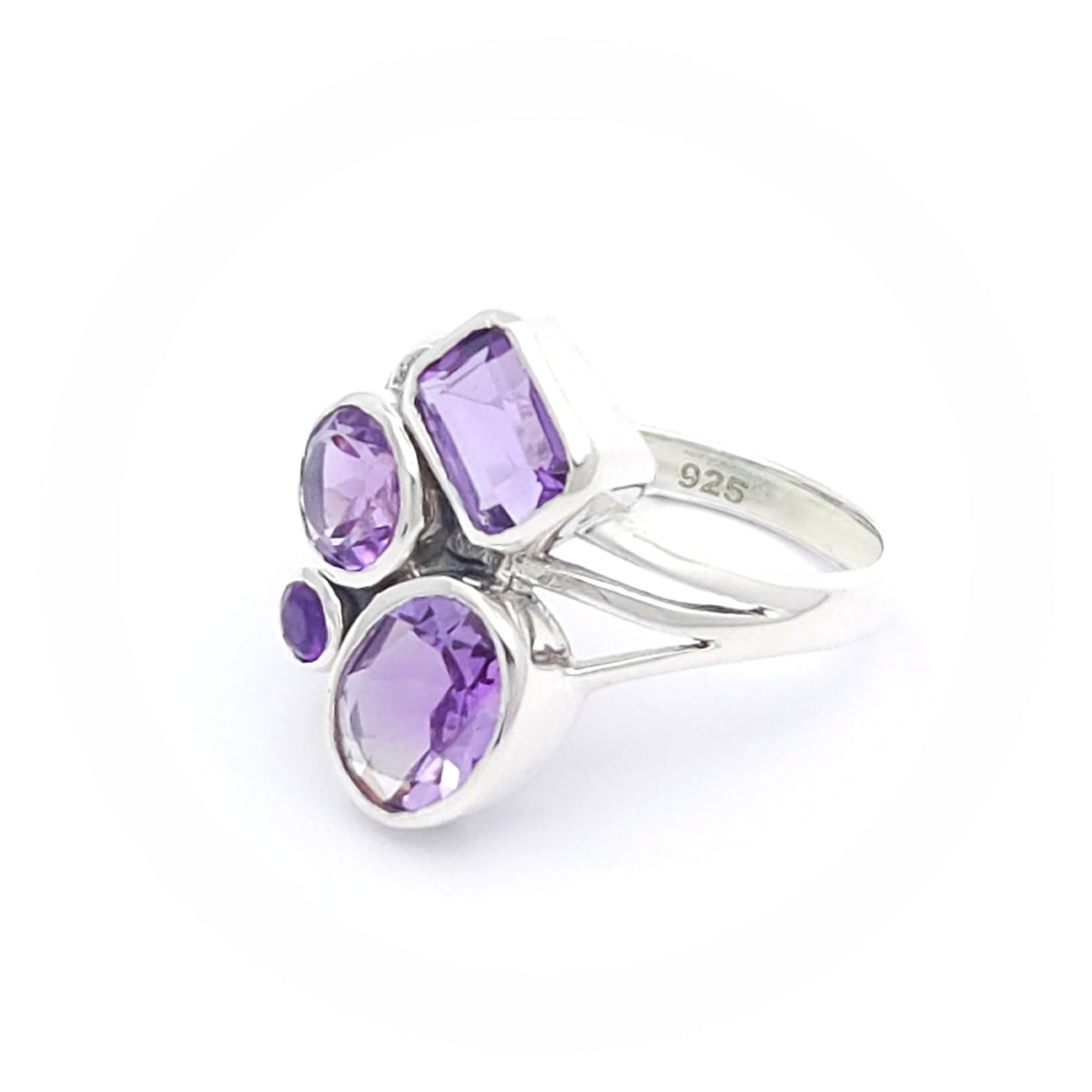 A silver ring featuring four amethyst gemstones of varying shapes and sizes. The gemstones include a rectangular cut, a round cut, an oval cut, and a small circular cut, all set in a modern, artistic design.