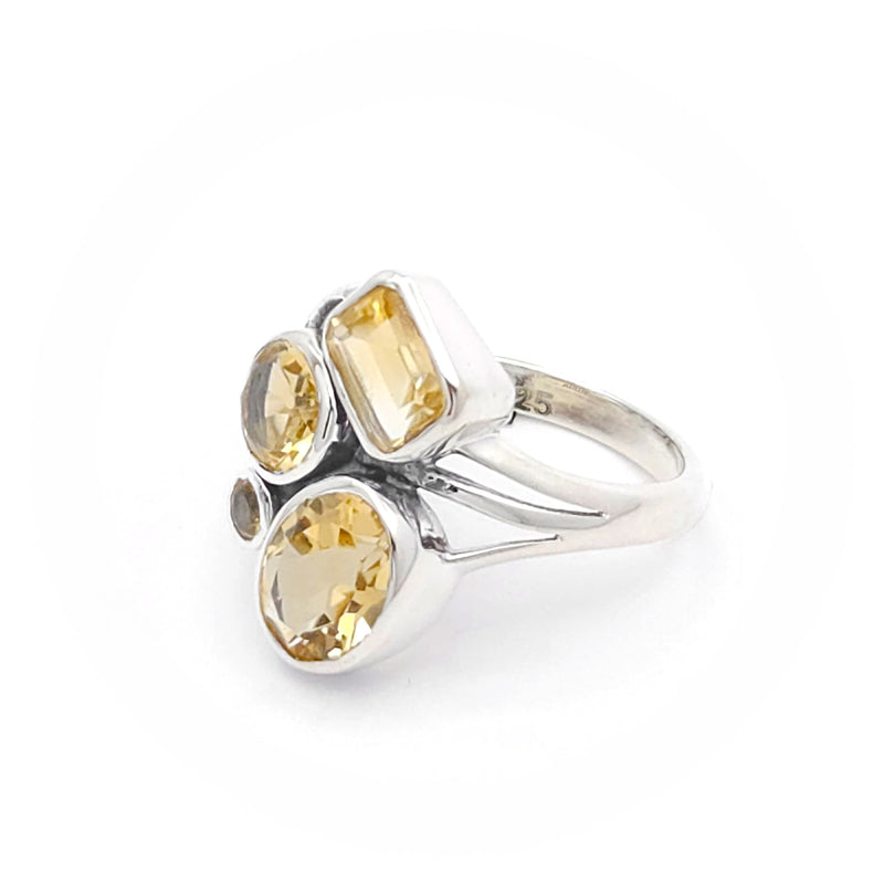 The image shows a silver ring adorned with four citrine gemstones. The gemstones are of different shapes: a rectangular cut, two round cuts, and an oval cut. The arrangement and variety of gemstone shapes make the ring visually interesting and unique.