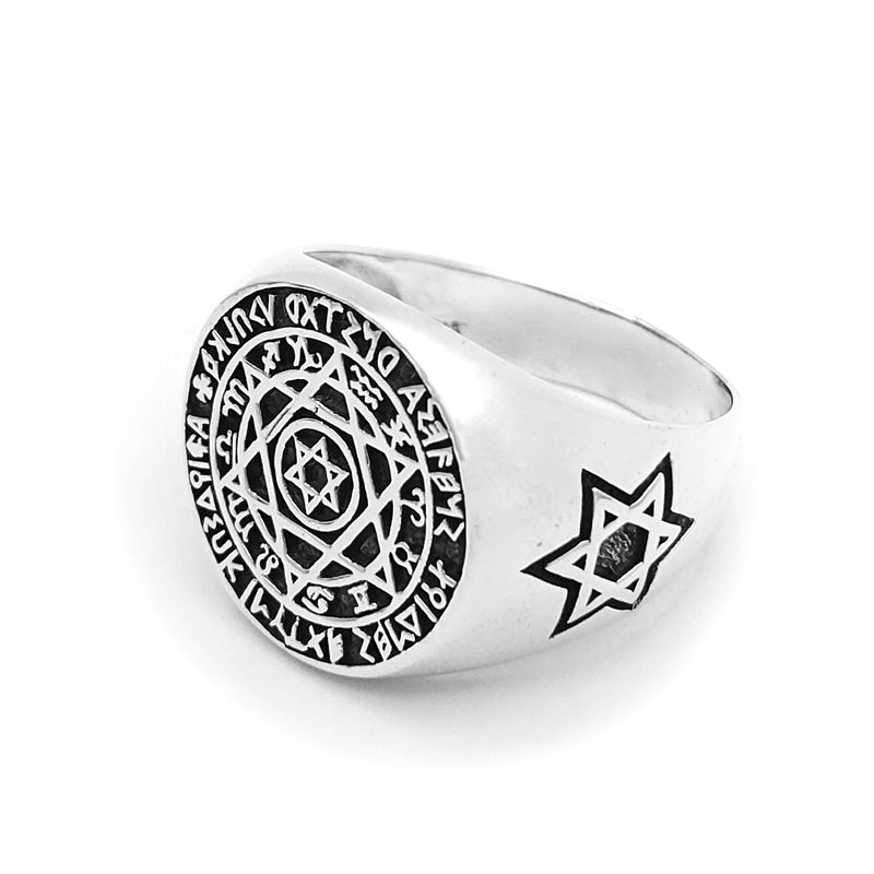 A silver ring featuring intricate designs and symbols. The central design is a hexagram (Star of David) surrounded by concentric circles filled with various symbols and characters. The outer circle contains a series of runic or mystical characters.